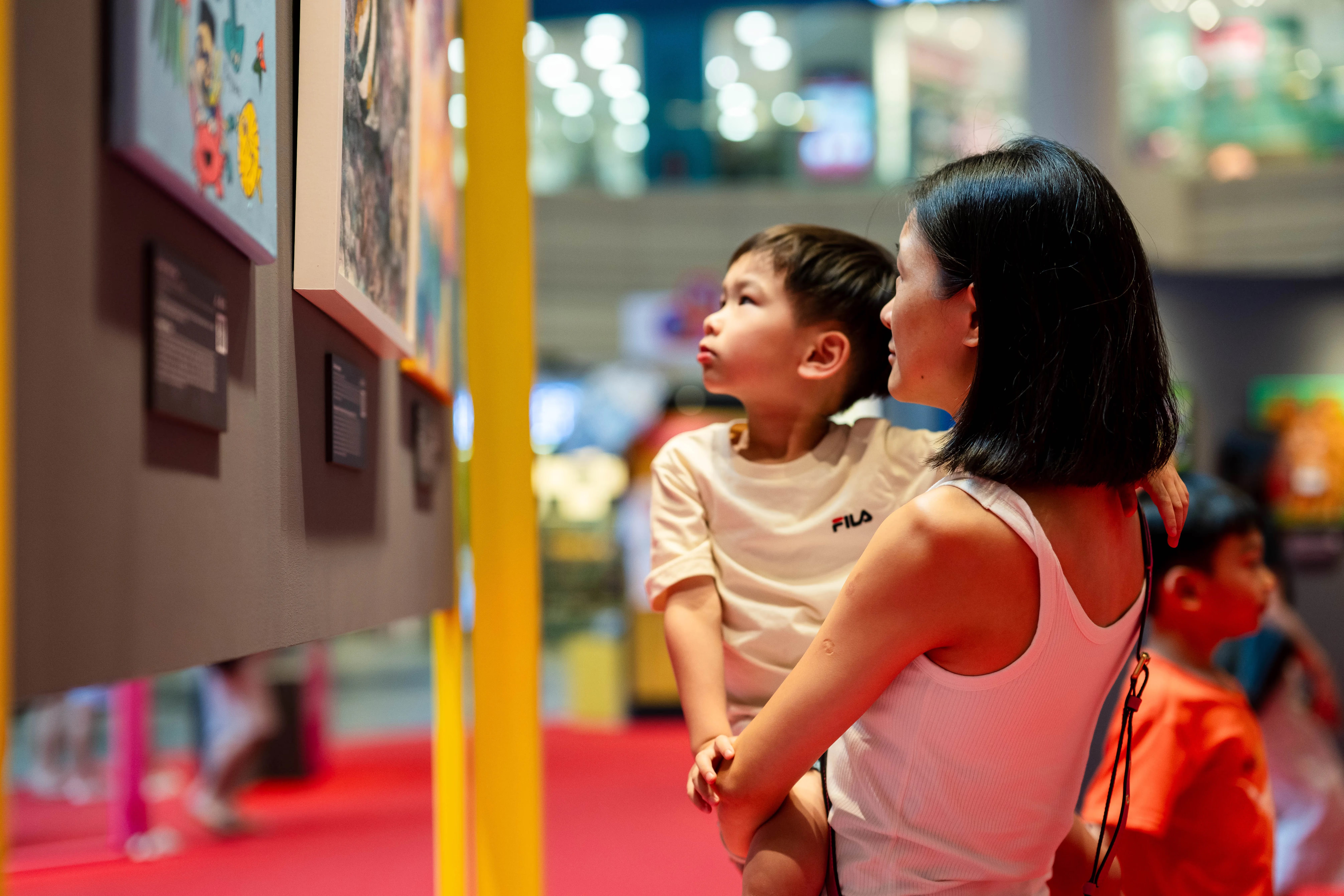 A woman holding a child is observing colorful artwork in a brightly lit gallery with red flooring.
